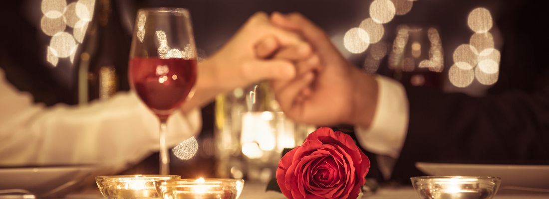 Un couple se tient la main entouré d'un verre de vin, d'une bougie parfumée et d'une rose rouge